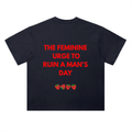 A black short-sleeve heavyweight cotton t-shirt shown from the back, featuring the phrase 'THE FEMININE URGE TO RUIN A MAN'S DAY' printed in bold red capital letters, with four small strawberry graphics underneath.