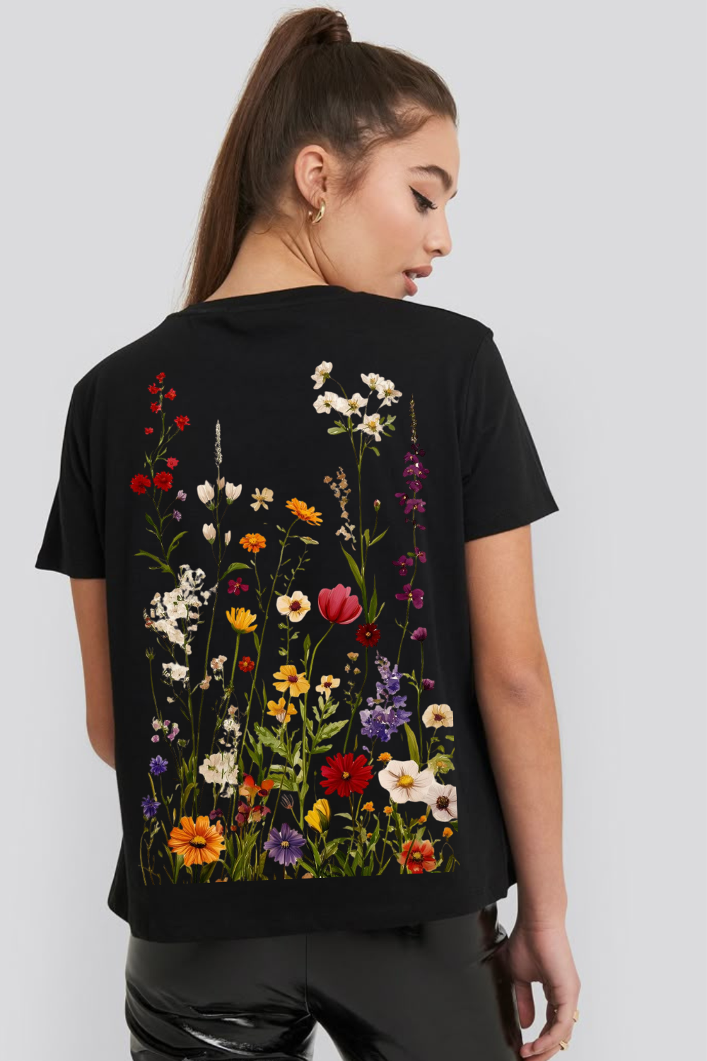 A woman shown from the back wearing a black short-sleeve T-shirt with a large colorful wildflower print across the back, paired with black pants, against a light gray studio background.